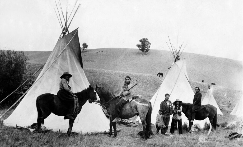 One b/w glass-plate negative and sepia photograph of several Native Americans (Ute) mounted on horseback and standing in front of teepees, circa 1900. One of the men is holding a gun. 1900- Image taken by James "Horsethief" Kelley.