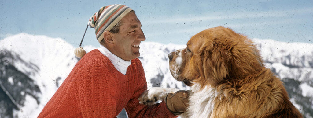 One color slide transparency of Fred Iselin and his St. Bernard named Mumbo, circa 1960. Fred is wearing a brightly colored striped hat and a red sweater.