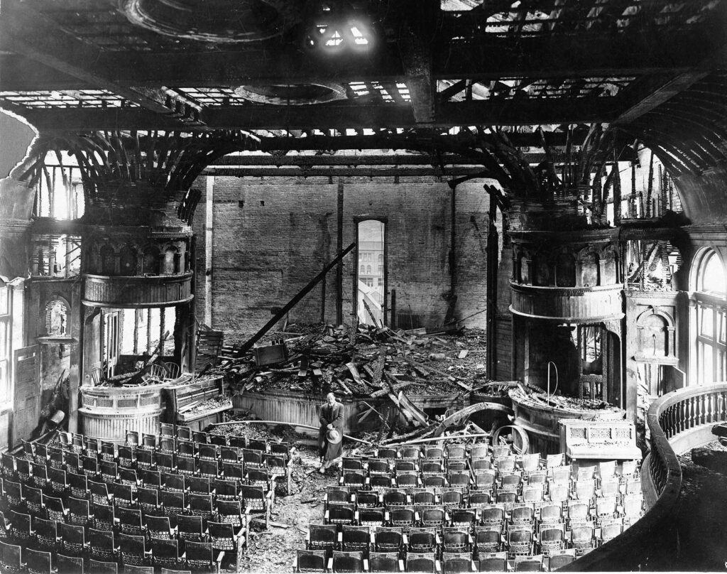 One 3.75" x 4.5" b/w photograph of the interior of the Wheeler Opera House after the fire of 1912. The interior is badly damaged and the roof of open to the sky. Through the back door/window, the Hotel Jerome is visible. There is a man standing in the middle of the picture.