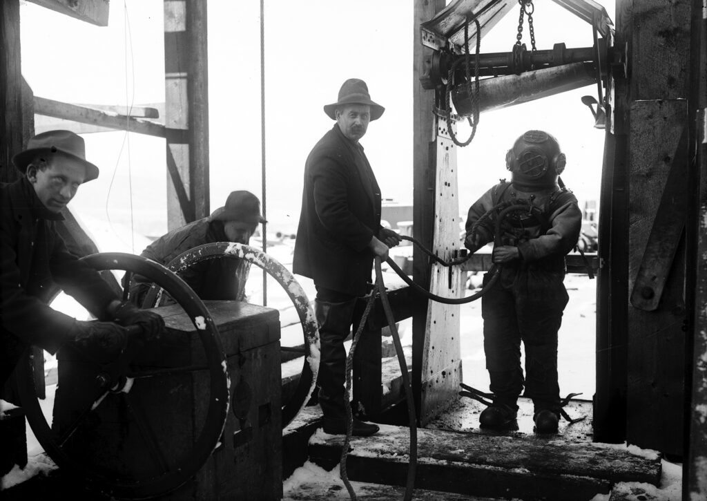 5x7 glass plate Negative and b/w photograph showing a deep sea diver (Fred Johnson) about to descend underground in the Smuggler Mine to renew the packing in a pump that had been drowned out. The man helping him is George Peterson, both of New York. The stalled pump was about 65 feet under water. The other two men identified in the paper as Jesse Yates far left and Joe Hull on the other side of the crank wheel. 1910 - Published on the front page of the November 30, 1910 Aspen Democrat-Times paper.