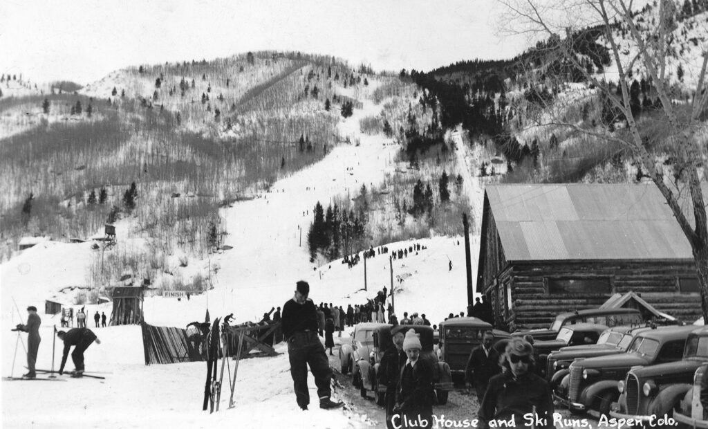 One glossy photo the 1941 National Championships on Aspen Mountain. The image shows the base of Aspen Mountain, the Corkscrew ski run, cars, people and a log structure. Written at the bottom of the postcard is "Club House and Ski Runs, Aspen, Colo.".