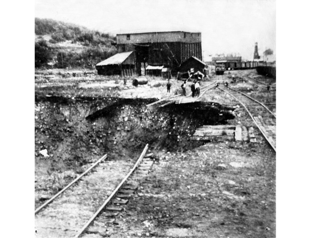 One mounted b/w photograph of the collapsed Glory Hole. Four men are standing on the tracks toward the back of the hole. The mine buildings are in the background. The collapse occurred in Saturday August 10th, 1918.