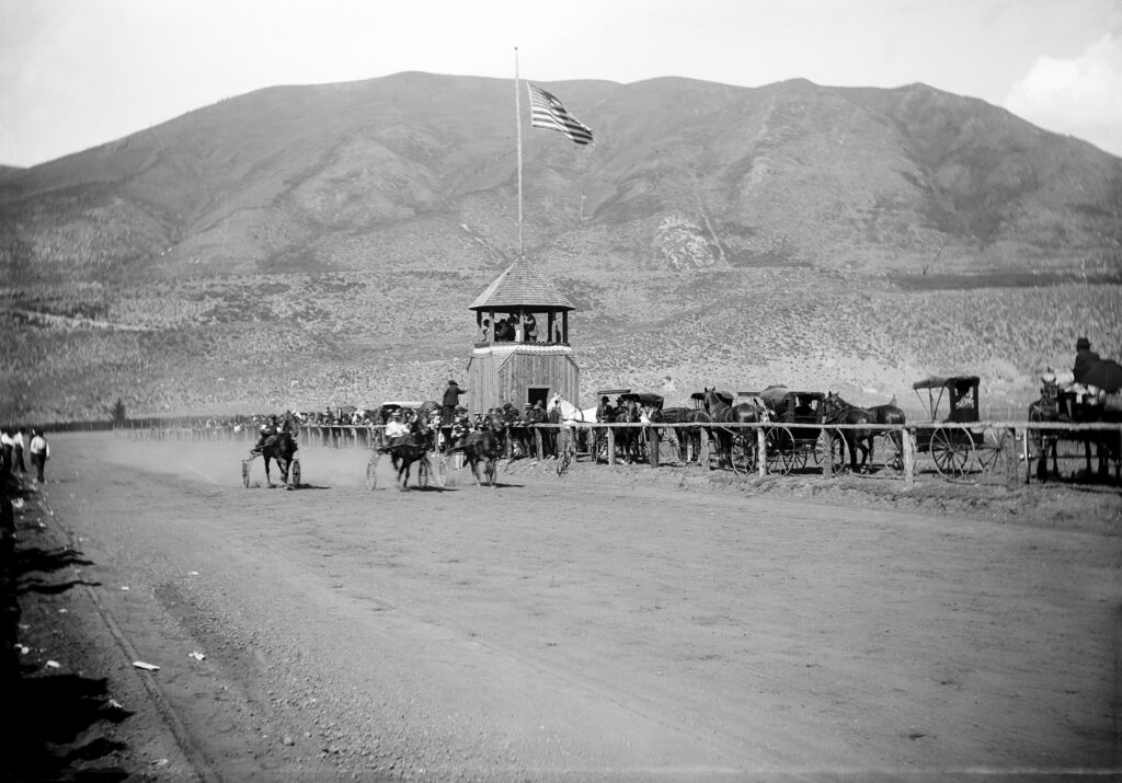 One b/w glass-plate negative of a horse race. There is the bandstand with the announcers and a flag on top, horse and buggies on side with the spectators as well as th horse drawn carts on the race track. Red Mountain is in the background. The horse track was in the west end where the Aspen Meadows is (2007).