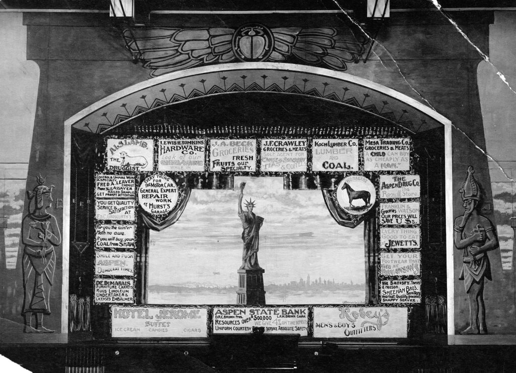 One b/w photograph of the interior of the Isis Theater, circa 1915. The photo shows the proscenium arch and the surrounding decoration. The curtain is down, showing advertisements for various Aspen businesses, and a painting of the Statue of Liberty in the center of the curtain. When the show was about to commence, the curtain would be raised.