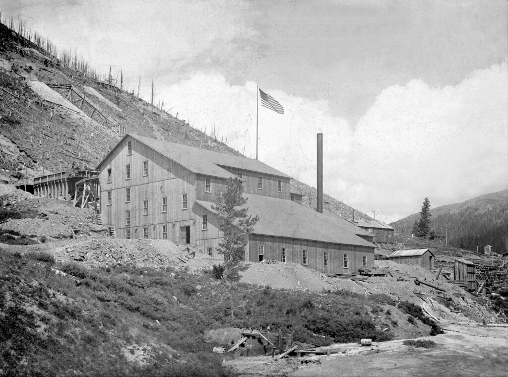 One mounted b/w photograph of the Farwell Mill at Independence, when it was still in use. The trees on the hillside have all been cut down, and there is an American flag flying from the top of the building. Date gathered from counting stars on flag. 1896-