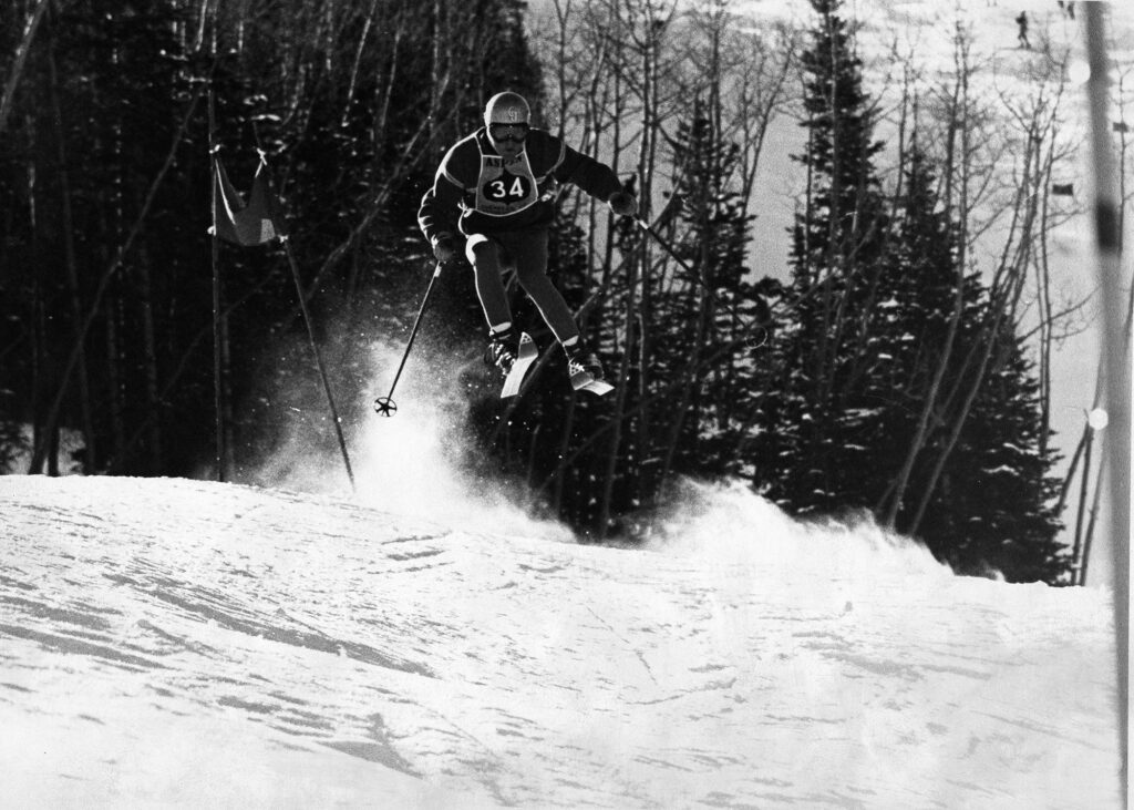One b/w glossy photograph of Spider Sabich in mid-air during a ski race. A gate can be seen behind him and he is wearing a bib that has the Aspen name and leaf, 1968-. Possibly the combined World Cup/Roch Cup. He placed 7th in the Men's DH that year.