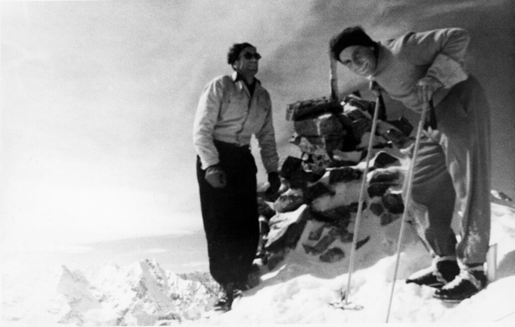 One b/w photograph of Andre Roch (left) and Billy Fiske (right) at the top of Mt. Hayden. Both men are in ski clothing and are standing by a pile of rocks. Behind them other mountain peaks can be seen, 1937.