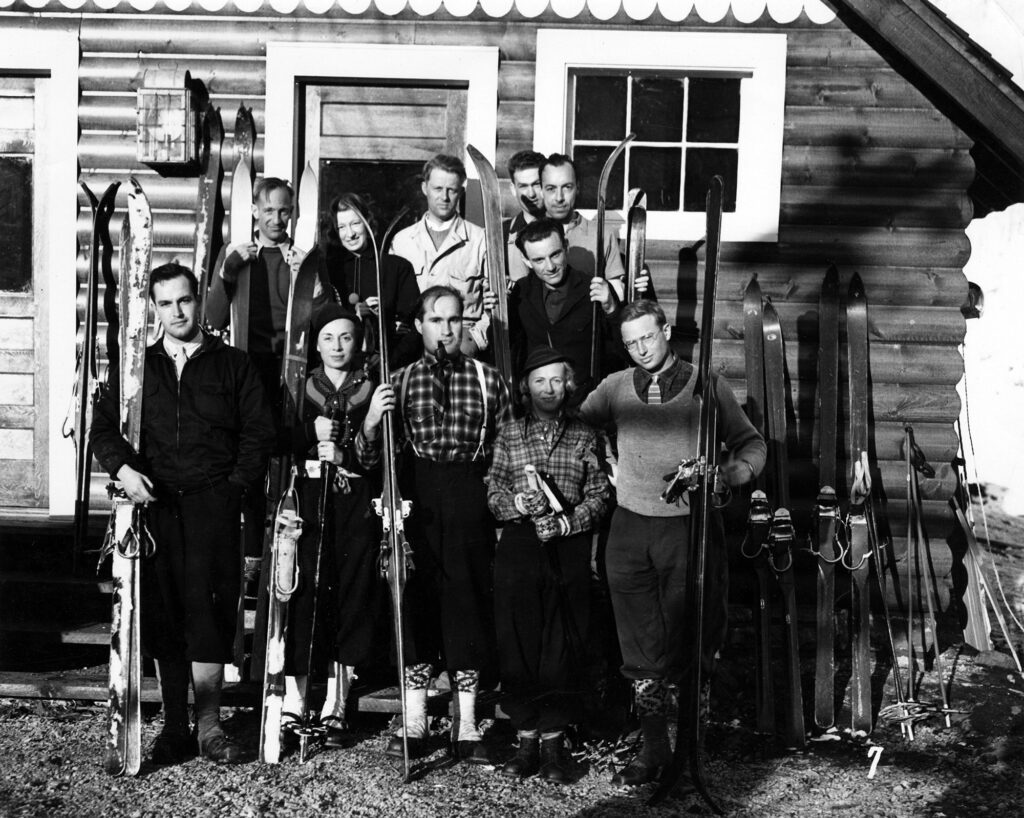 One b/w photograph of eleven people lined up on the front steps of the Highland Bavarian Lodge holding skis, 1936. Five people are standing on the ground in front of the steps, and there are six people standing on the steps. There are two pairs of skis and poles just to the right of the group leaning against the lodge. Pictured in the top row are Andre Roch, Gretl Arnold Fuler, Steve Hart, Norman Barwise, and an unknown (possibly James S. Bodrero) Italian Consul. In the bottom row is William V. Hodges, Martha Wilcox, Joseph Hodges, Polly Grimes, and Frank Ashley. December 1936.