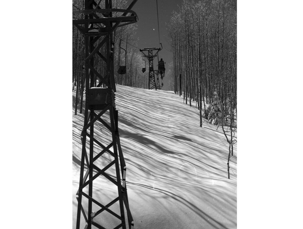 One b/w negative of skiers riding up Lift One, circa 1960. The canvas blankets used to keep people warm on the ride up are visible on the empty chairs coming down. There is untracked powder below the lift, and there are aspen trees on either side of the lift line.