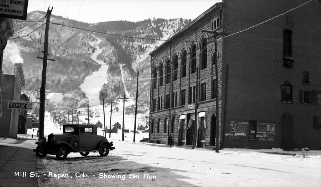 One film negative of the Wheeler Opera House from Mill Street facing Aspen Mountain, 1939. Roch Run and Corkscrew can be seen in the background, and there is a car on the left side of the image. This image was made into a postcard that was sold at the Cooper Book and Stationery Company; the caption reads "Mill St.- Aspen Colo.- Showing Ski Run."
