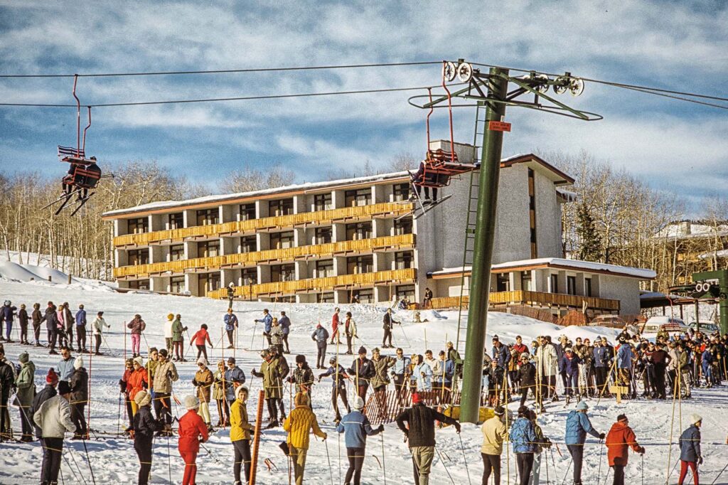 One color digital print of Fanny Hill with part of the lower chairlift tower as well as the Burlingame Lift at Snowmass-at-Aspen, 1968. There is a line of people waiting to load the lift from the top of the Fanny Hill Lift. The Silvertree Inn is the in background.