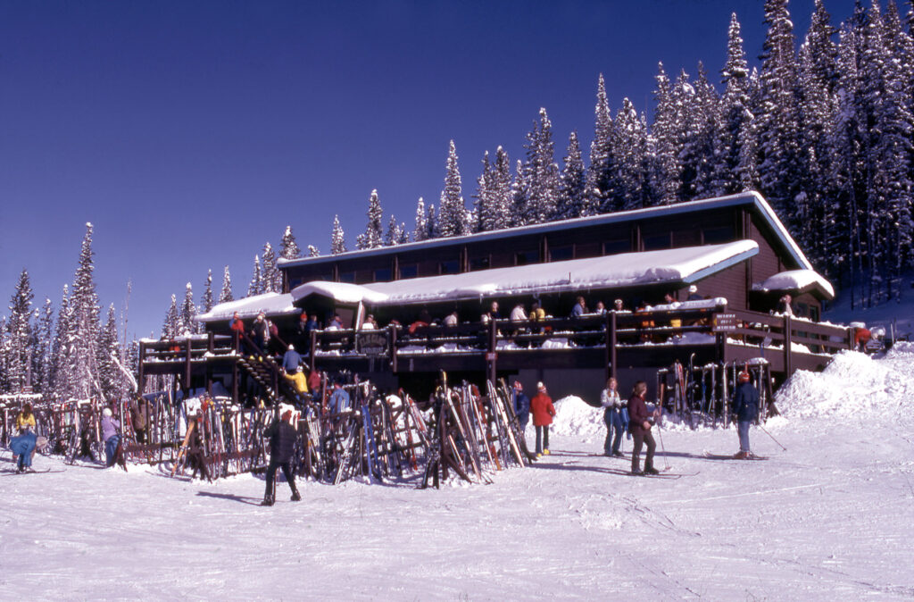 One color slide of skiers in front of the Elk Camp Restaurant at Snowmass, 1975. The ski racks are filled with skis, and there are people on the deck. This became the Cafe Suzanne in 1989, and the building was torn down in 2012 when a new Elk Camp Restaurant was built at the top of the nearby Elk Camp Gondola (built in 2006).