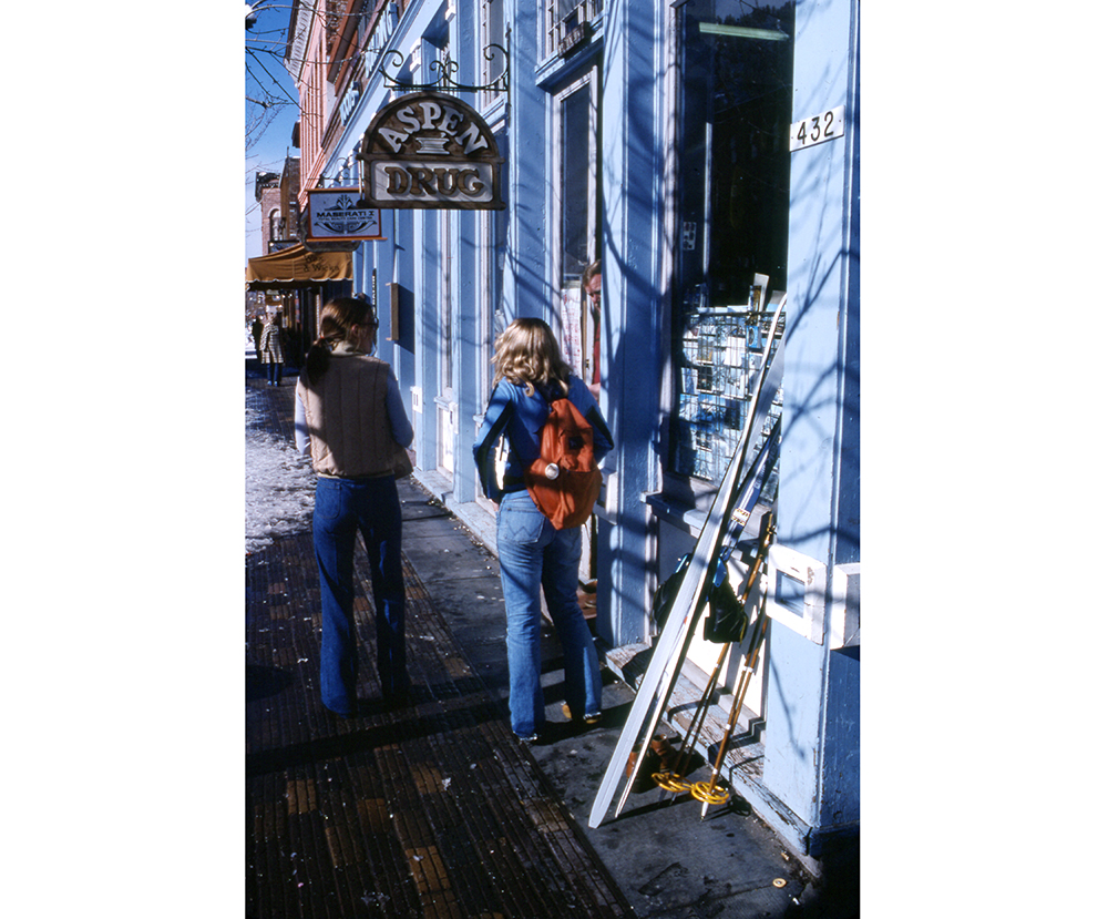 One color slide of two women standing in front of the Woods Building on E. Hyman Avenue, 1978. A sign for Aspen Drug is above their heads, and there is a pair of skis leaning against the building. The awning for Wax and Wicks can be seen down the street.