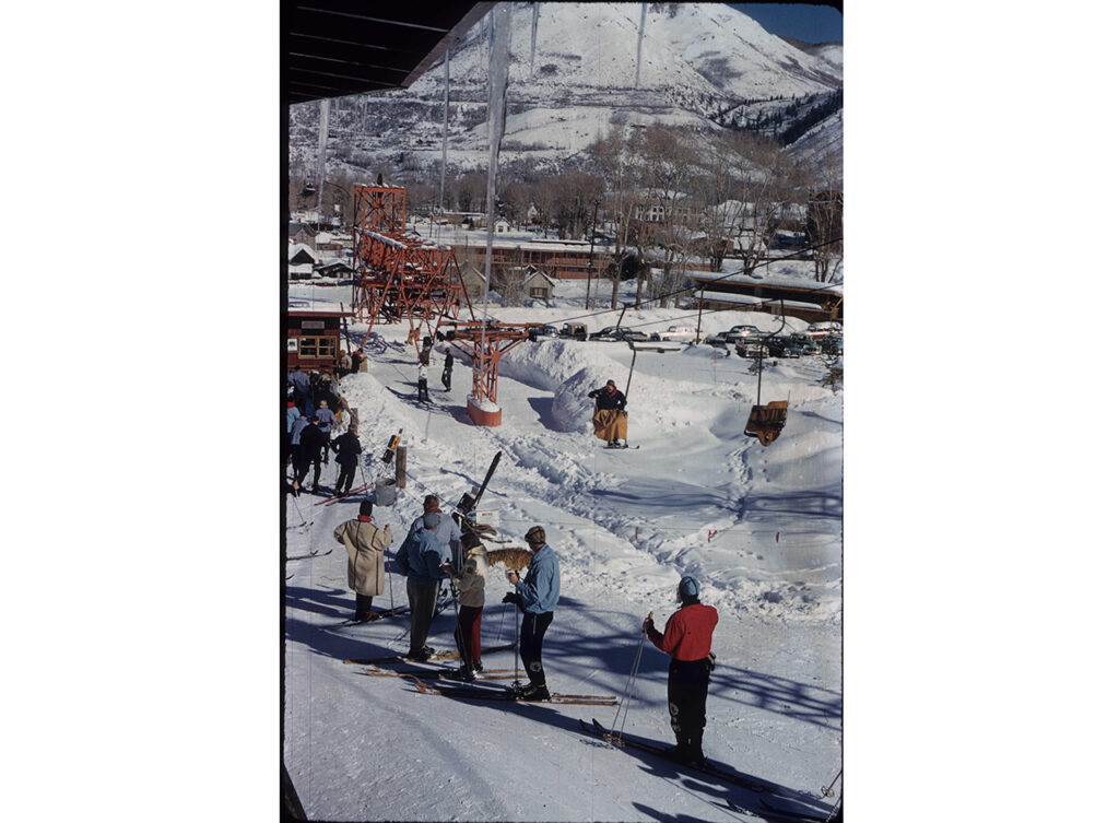 One color slide transparency of skiers lined up at the base of Aspen Mountain waiting to get onto Lift One. People riding the lift have the canvas blanket over them. Buildings around town can be seen in the background, circa 1955.